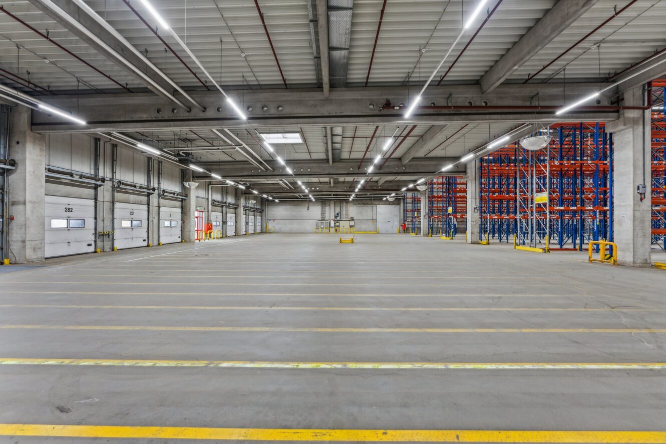 Geräumiges, leeres Lagerhaus mit Betonböden, hohen Decken, Industriebeleuchtung, Laderampe und leeren Regalen an der rechten Wand.