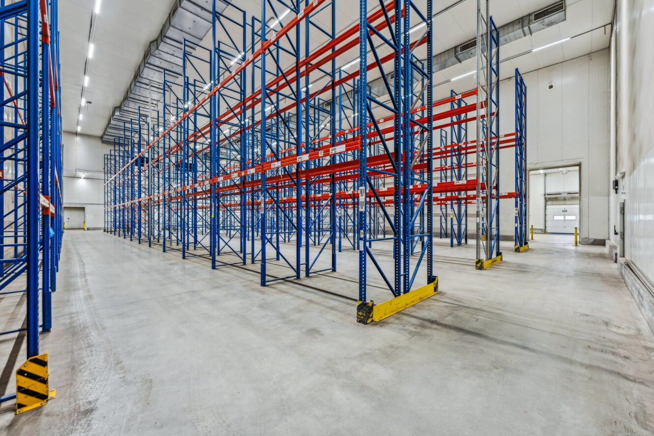 Leeres Lagerhaus mit hohen blauen und roten Metallregalen, Betonboden, hoher Decke und heller Beleuchtung.
