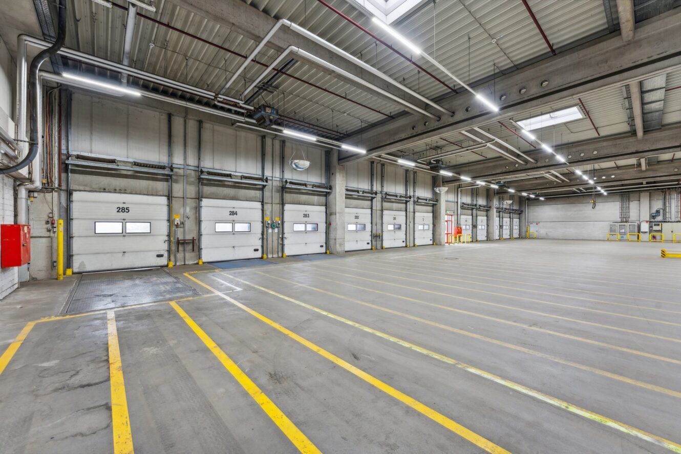 Geräumiges, leeres Industrielager mit mehreren Laderampen, markierten gelben Linien auf dem Boden und Oberlichtern an einer Metalldecke.