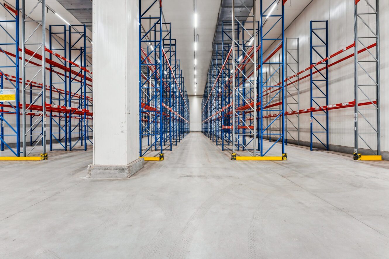 Ein leeres Lagerhaus mit hohen blauen und roten Metallregalen, weißen Wänden und heller Deckenbeleuchtung.