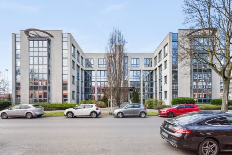 Modernes, mehrstöckiges Bürogebäude mit großen Fenstern und zentralem Eingang, vor dem die Autos entlang der Straße geparkt sind, mit Bäumen auf beiden Seiten.