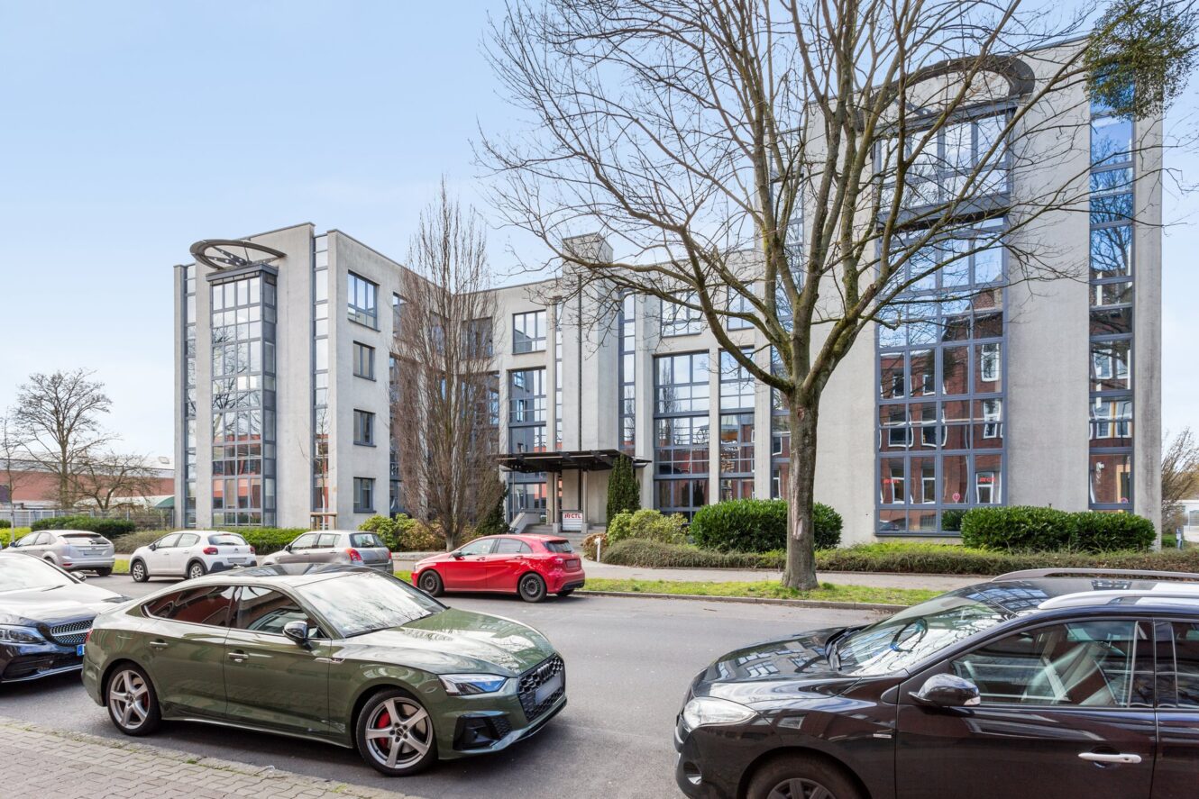 Mehrere geparkte Autos säumen eine Straße vor einem modernen, mehrstöckigen Bürogebäude mit großen Fenstern und Bäumen in der Nähe.