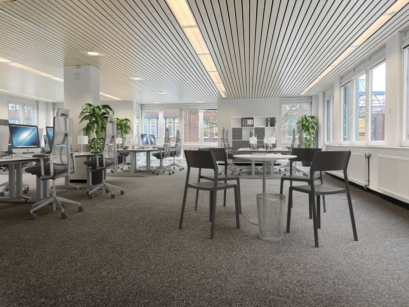 Modernes Großraumbüro mit Schreibtischen, Computern, Stühlen und Pflanzen. Im Vordergrund steht ein runder Tisch mit vier Stühlen in der Nähe großer Fenster. Der Raum ist durch natürliches Licht gut beleuchtet.
