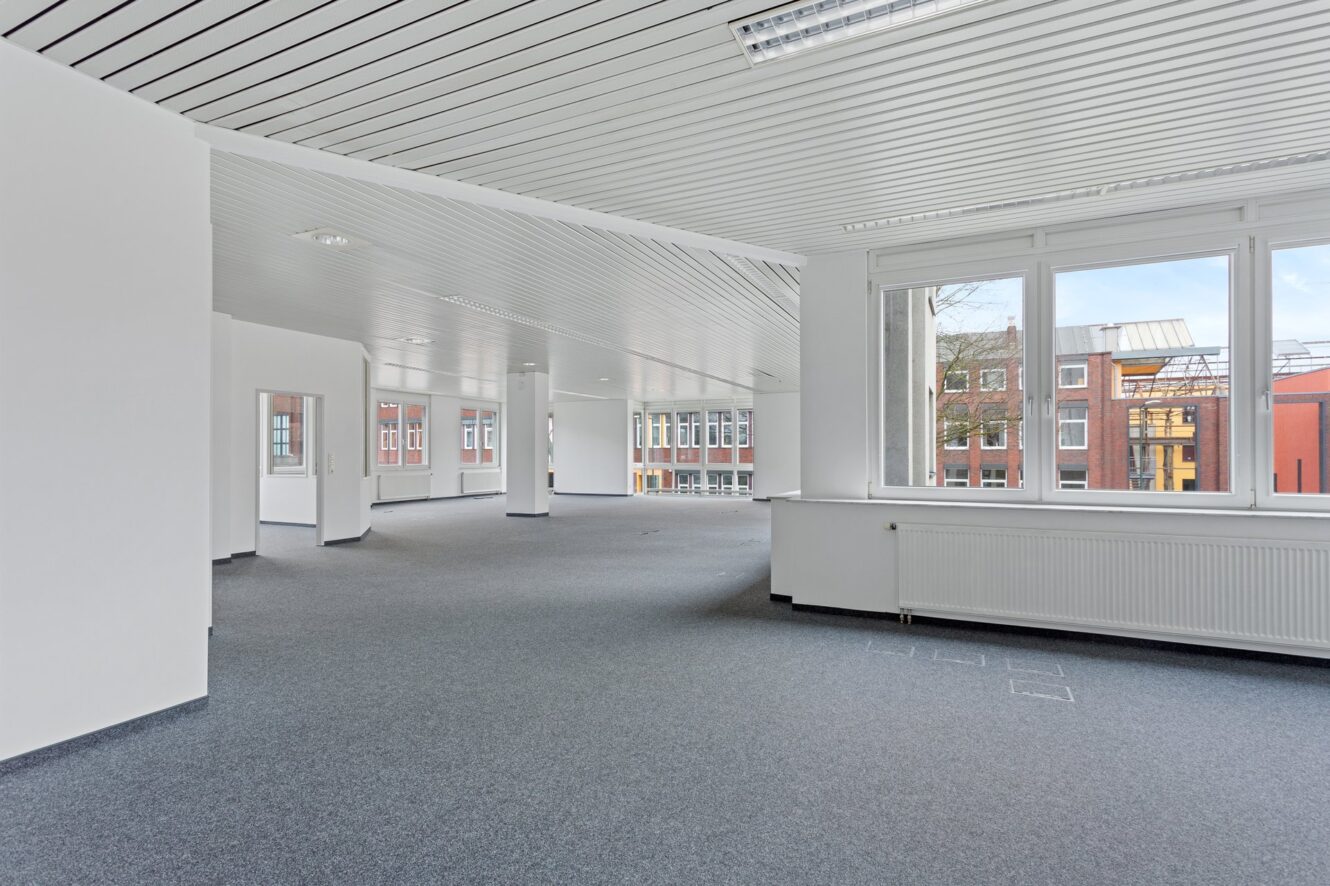 Leere, moderne Büroräume mit weißen Wänden, großen Fenstern, grauem Teppichboden und Deckenleuchten, die durch die Fenster einen Blick auf die umliegenden Gebäude bieten.