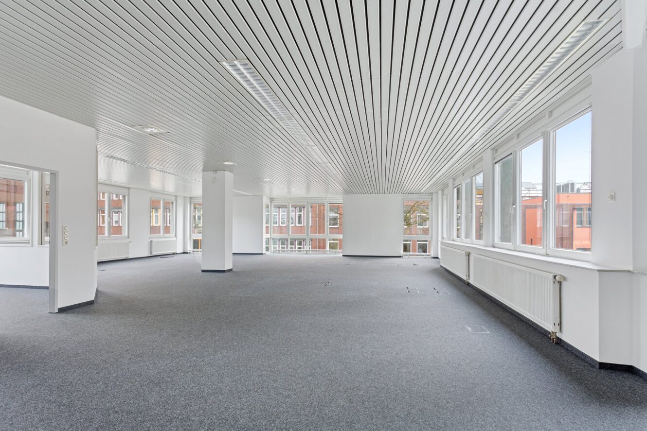Ein großer, leerer Büroraum mit grauem Teppich, weißen Wänden, einer gestreiften Decke und großen Fenstern, die für Tageslicht sorgen.