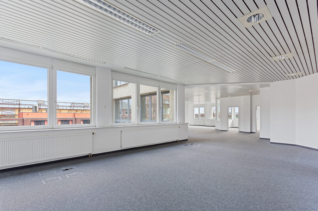 Leere, moderne Büroräume mit großen Fenstern, weißen Wänden und grauem Teppichboden, durch deren Fenster man die Außenanlagen sehen kann.