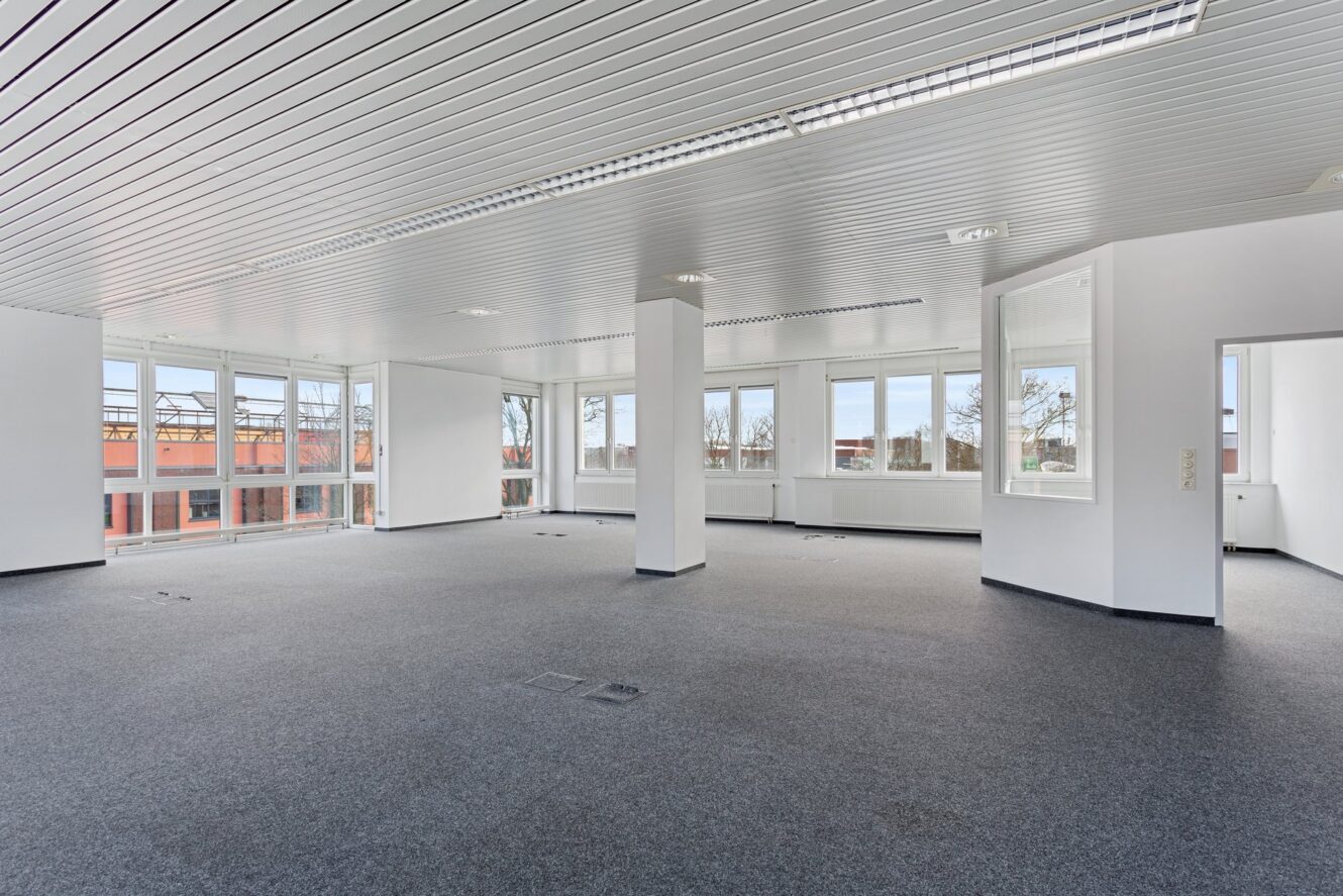 Leere, moderne Büroräume mit großen Fenstern, weißen Wänden, Teppichboden und Deckenleuchten.