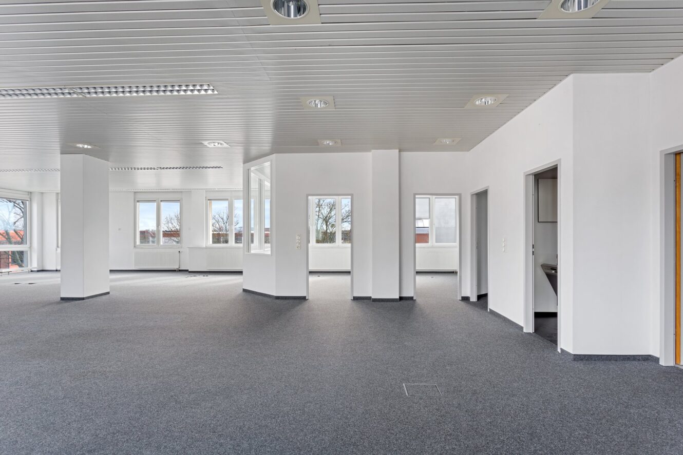 Geräumiges, leeres Büro mit grauem Teppich, weißen Wänden, mehreren Fenstern und mehreren offenen Türen. Durch die Fenster fällt natürliches Licht ein.