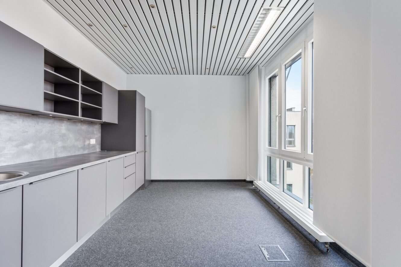 Moderne, unmöblierte Küchenzeile mit grauen Schränken, offenen Regalen, einer Spüle und großen Fenstern, die Tageslicht hereinlassen.