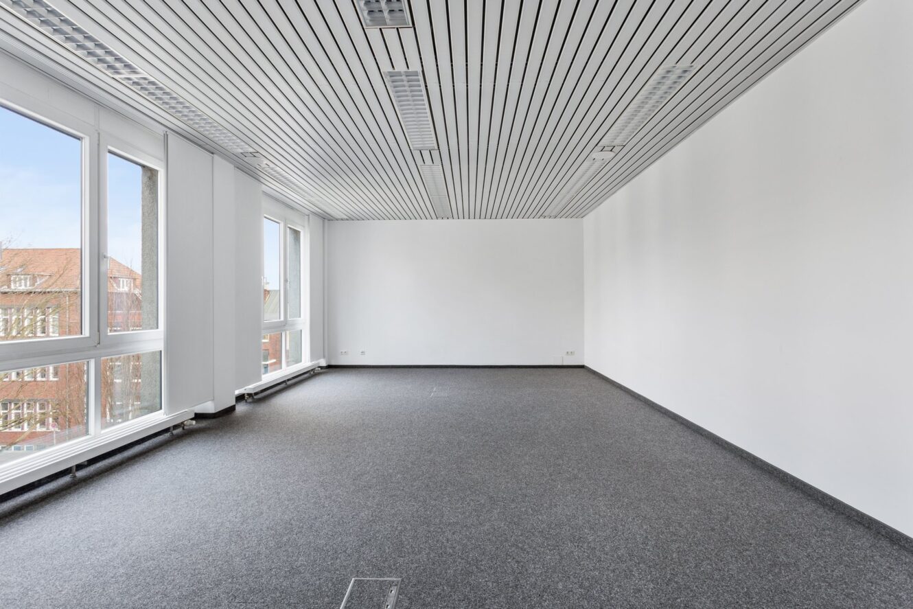 Ein leerer Büroraum mit grauem Teppich, weißen Wänden, großen Fenstern auf der linken Seite und einer weißen Lamellendecke mit Leuchtstoffröhren.