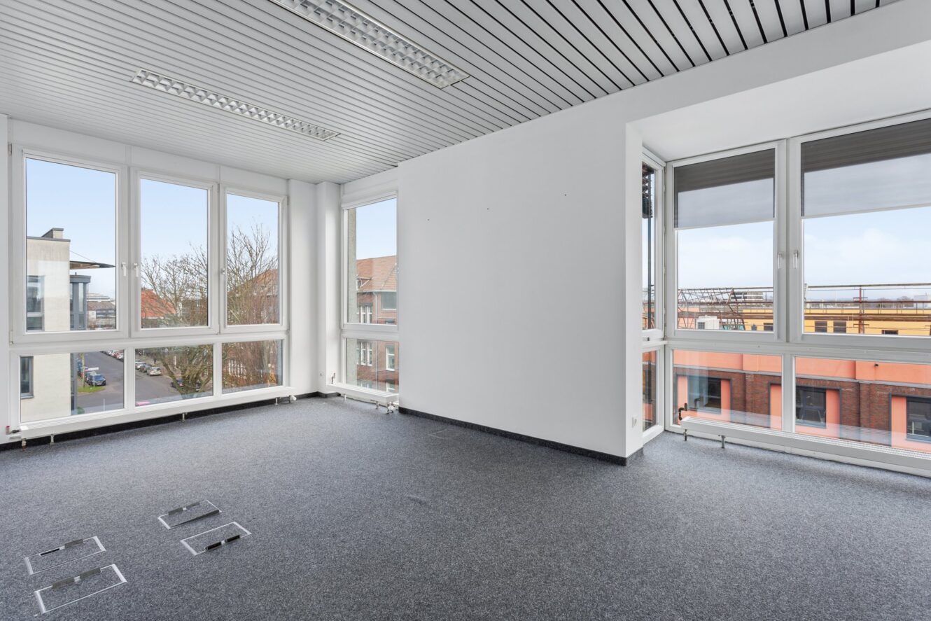 Leere Büroräume mit großen Fenstern, weißen Wänden, grauem Teppichboden und Leuchtstoffröhrenbeleuchtung. Städtische Gebäude und Straße durch die Fenster sichtbar.