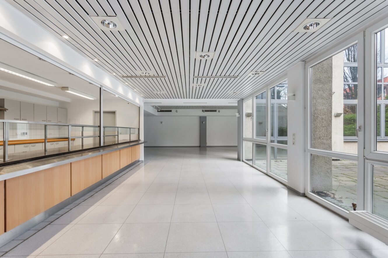 Große, leere Cafeteria mit gefliesten Böden, einer Ausgabetheke auf der linken Seite und raumhohen Fenstern auf der rechten Seite, durch die Tageslicht einfällt.