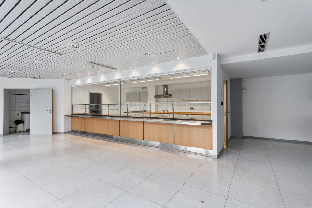 Geräumiger, leerer Raum mit gefliestem Boden, großen Fenstern und einer offenen Serviertheke mit Blick auf eine moderne Küchenzeile im Hintergrund.