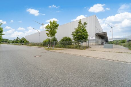 Eine große Industriehalle aus Metall, umgeben von einem Zaun und Bäumen, steht an einer leeren Straße unter einem teilweise bewölkten Himmel. Im Hintergrund sind Windkraftanlagen zu sehen.