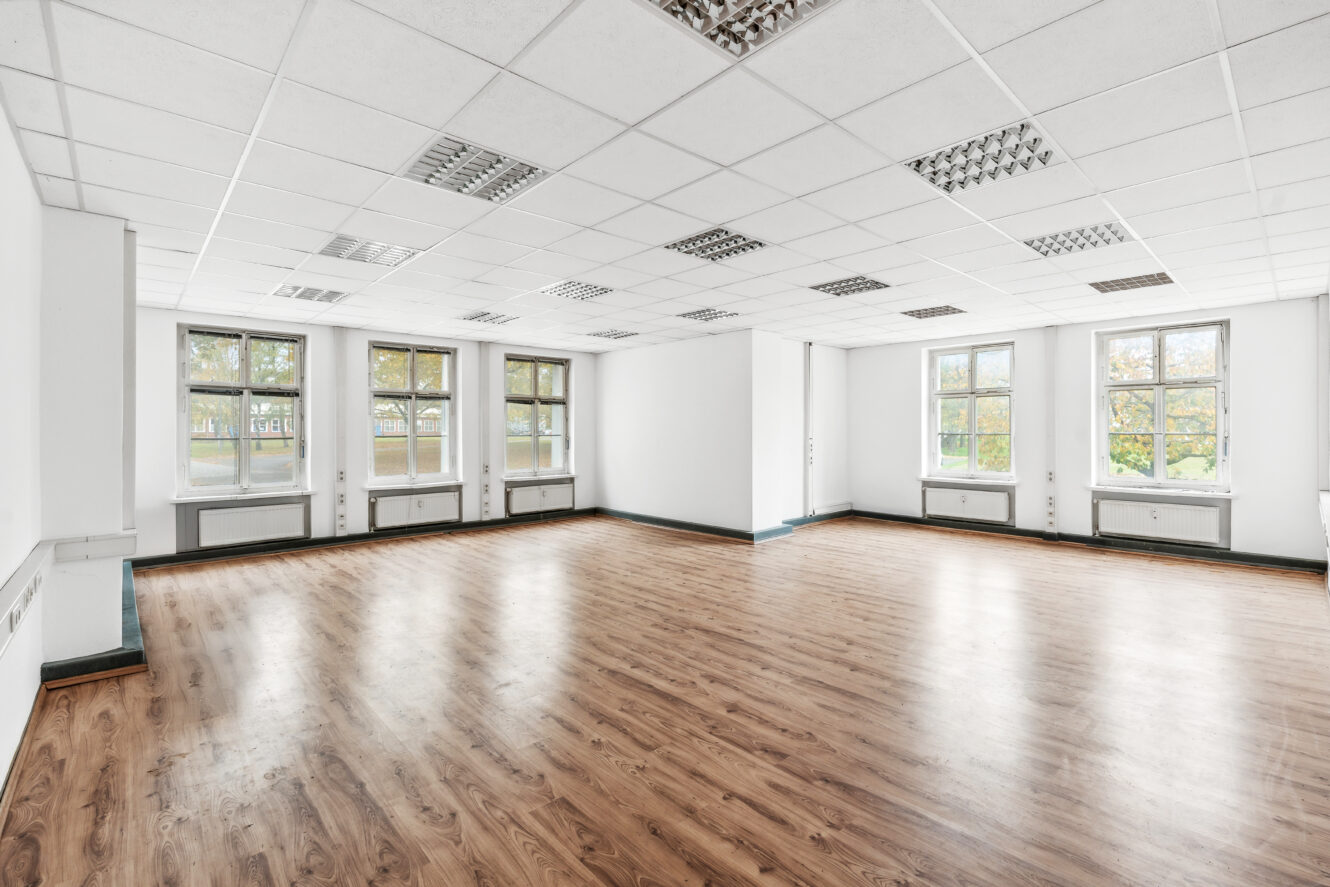 Leerer Büroraum mit weißen Wänden, großen Fenstern, Holzfußboden und fluoreszierenden Deckenleuchten.