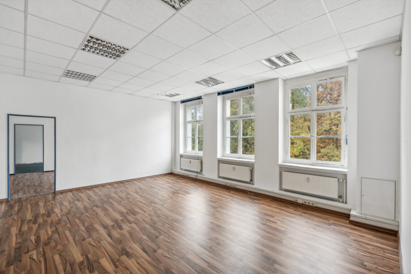 Ein leerer Raum mit weißen Wänden, großen Fenstern, die natürliches Licht hereinlassen, einem holzgemusterten Boden und einem Wandspiegel, der einen Teil des Raumes reflektiert.