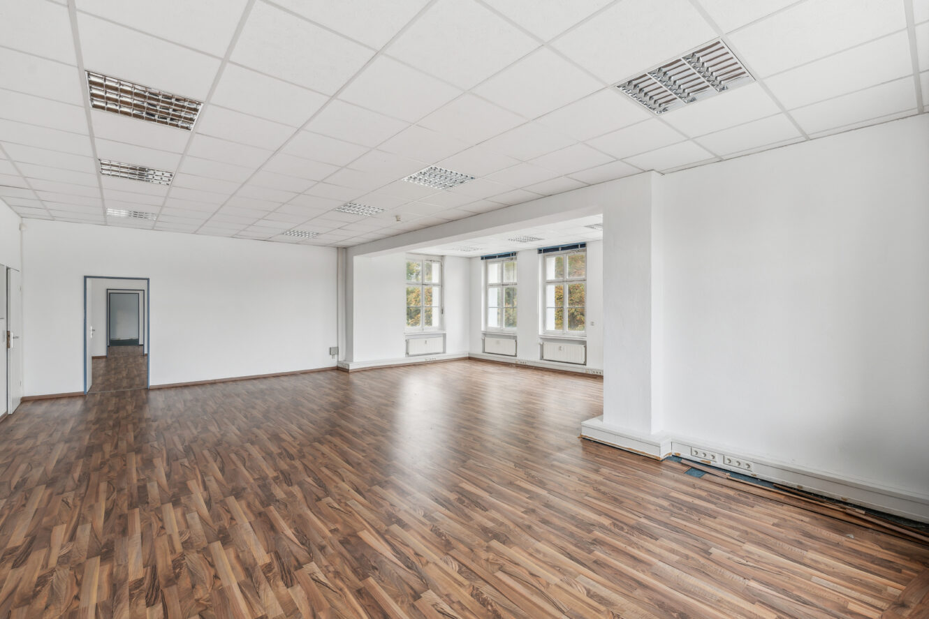 Geräumiger, leerer Raum mit weißen Wänden, großen Fenstern, holzgemustertem Fußboden und einer abgehängten Decke mit Leuchtstoffröhren.