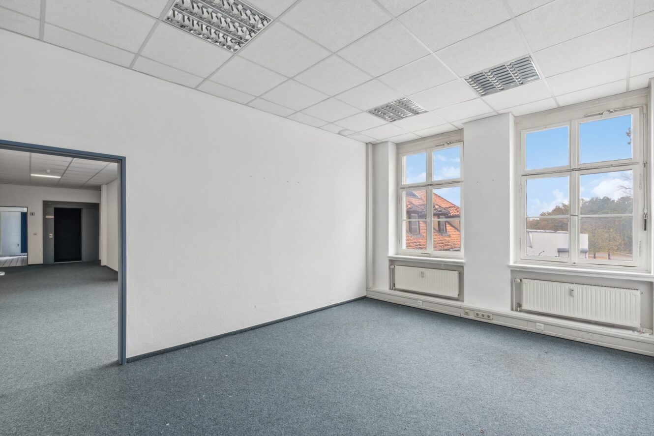 Leerer Büroraum mit weißen Wänden, blaugrauem Teppich, großen Fenstern und Leuchtstoffröhren an der Decke, angrenzend an einen anderen offenen Büroraum.