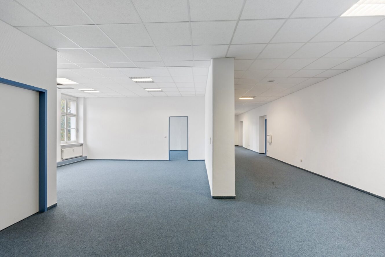 Leerer Büroraum mit blauem Teppich, weißen Wänden und einer Trennwand, die den Raum unterteilt; mehrere Türen und Fenster sind sichtbar.