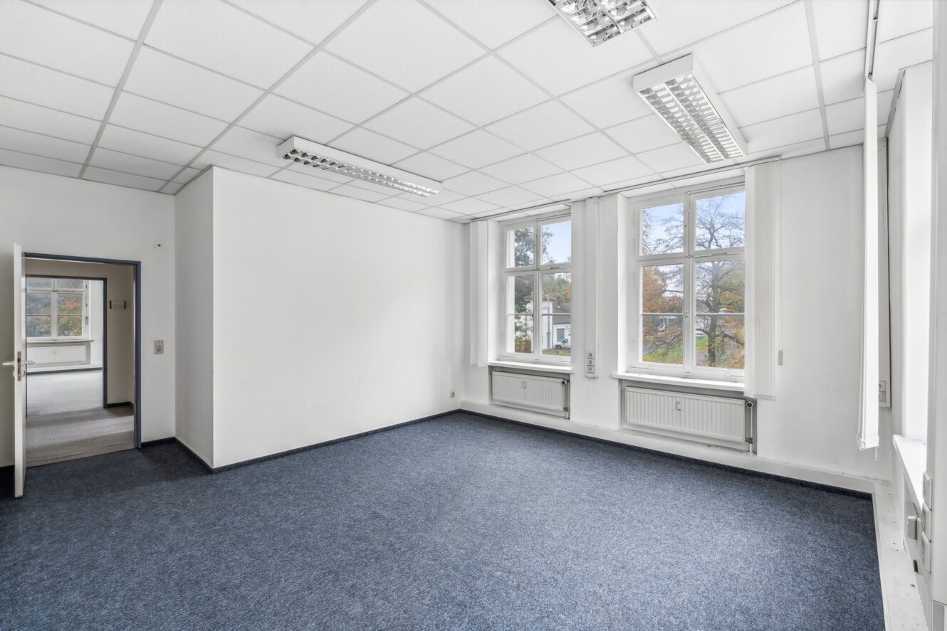 Ein leerer Büroraum mit weißen Wänden, blauem Teppich, zwei großen Fenstern, Deckenleuchten und einer offenen Tür, die in einen anderen Raum führt.