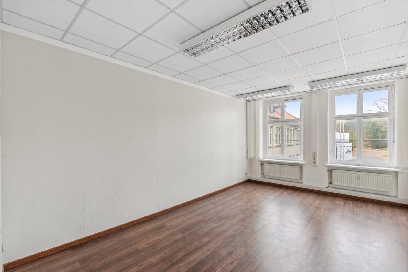 Leerer, unmöblierter Raum mit weißen Wänden, abgehängter Decke mit Leuchtstoffröhren, großen Fenstern und Laminatfußboden.
