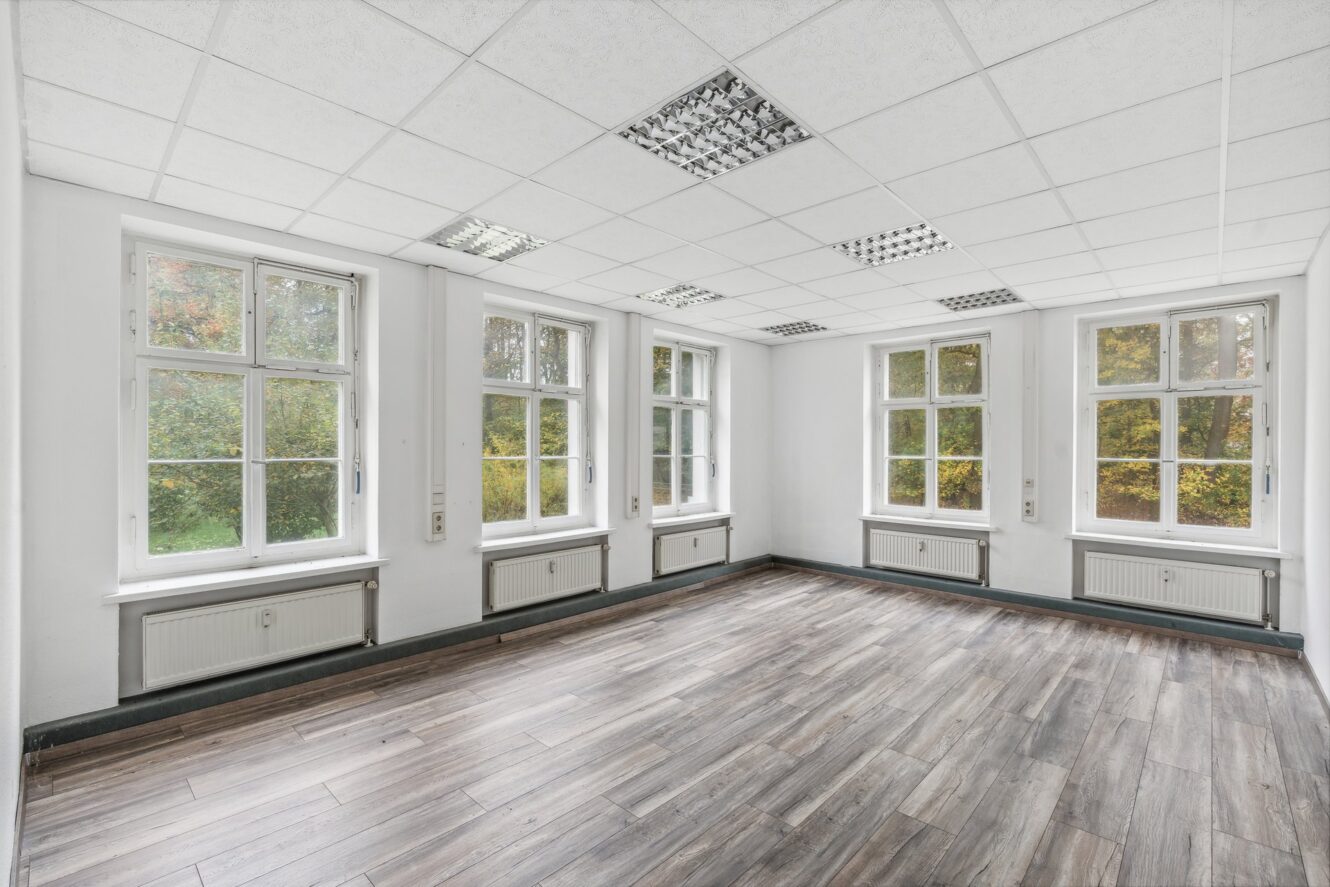 Ein leerer, heller Raum mit weißen Wänden, großen Fenstern, einer abgehängten Decke mit Leuchtstoffröhren und einem holzgemusterten Fußboden.