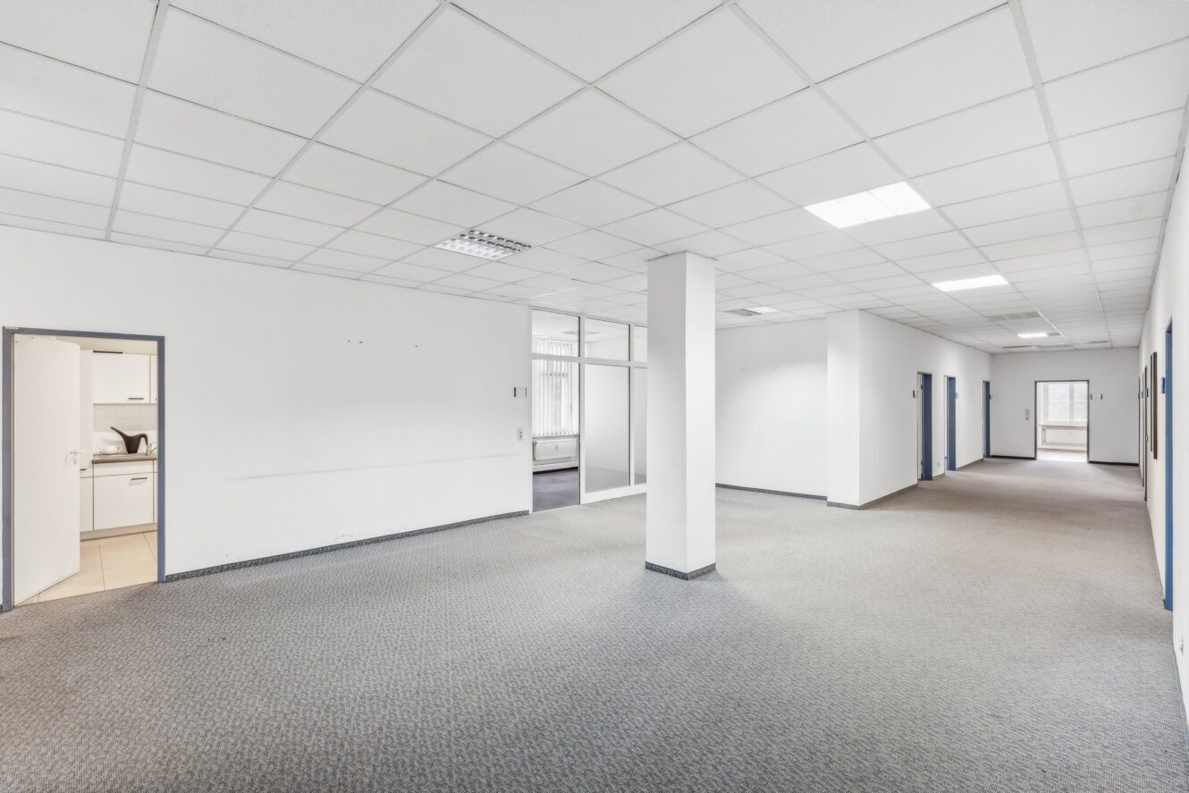 Geräumiges, leeres Büro mit Teppichboden, weißen Wänden, fluoreszierenden Deckenleuchten, einer zentralen Säule, einer Küchenzeile auf der linken Seite und gläsernen Trennwänden im Hintergrund.