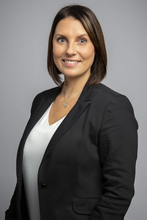 Eine Frau mit glattem braunem Haar, die einen schwarzen Blazer über einem weißen Oberteil trägt, steht vor einem schlichten grauen Hintergrund und lächelt in die Kamera.