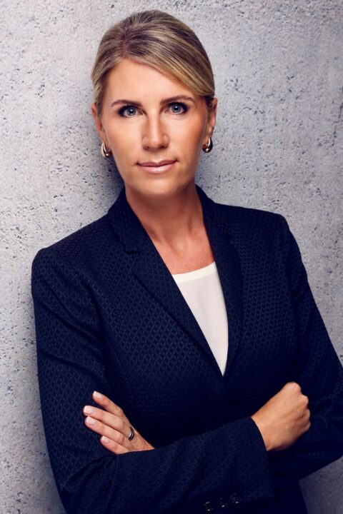 Eine Frau mit blondem Haar und tiefem Pferdeschwanz, die einen dunkel strukturierten Blazer und ein weißes Oberteil trägt, steht mit verschränkten Armen vor einer grau strukturierten Wand und schaut in die Kamera.