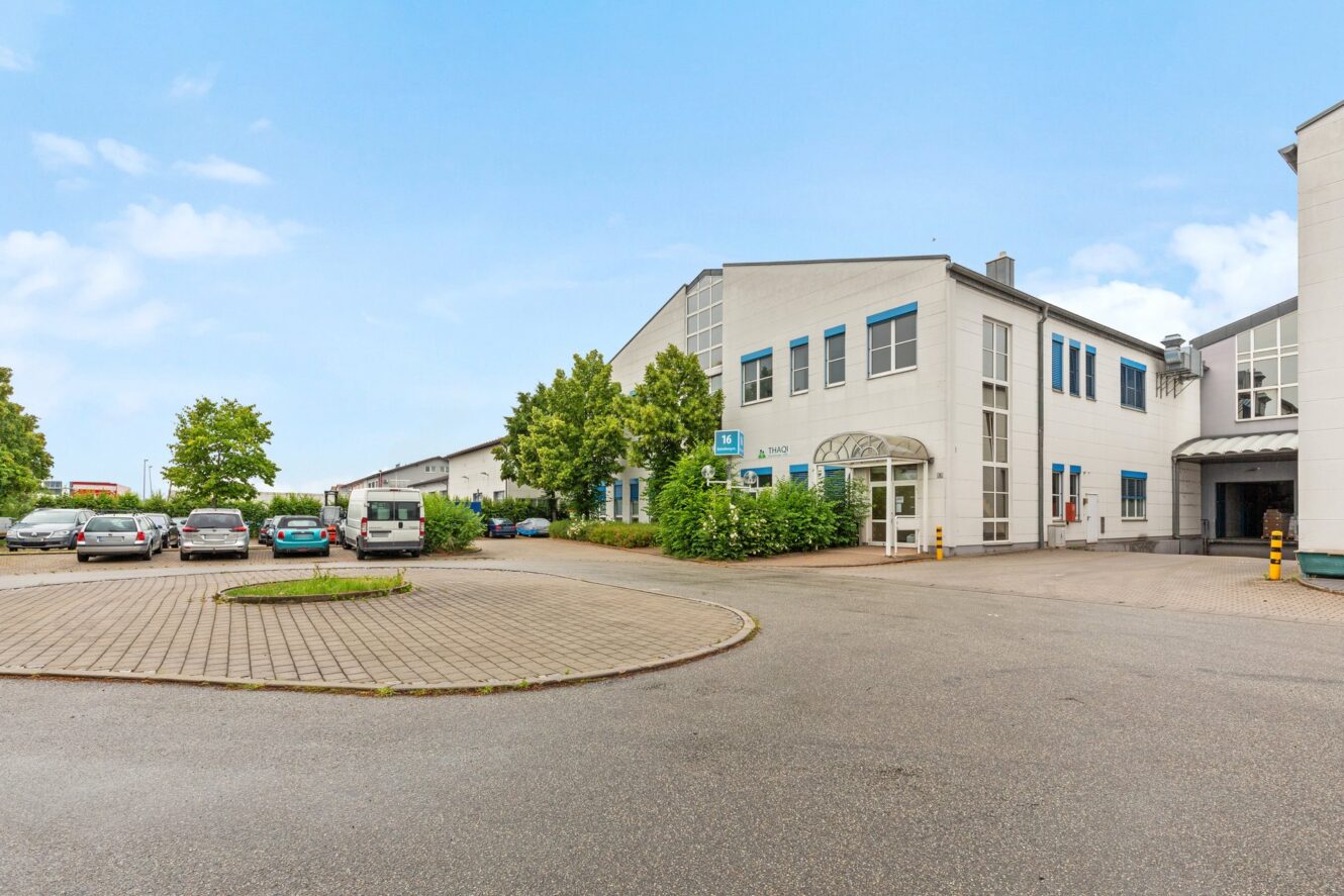 Ein modernes Industriegebäude mit großen Fenstern, umgeben von parkenden Autos und Grünanlagen, unter einem klaren Himmel.