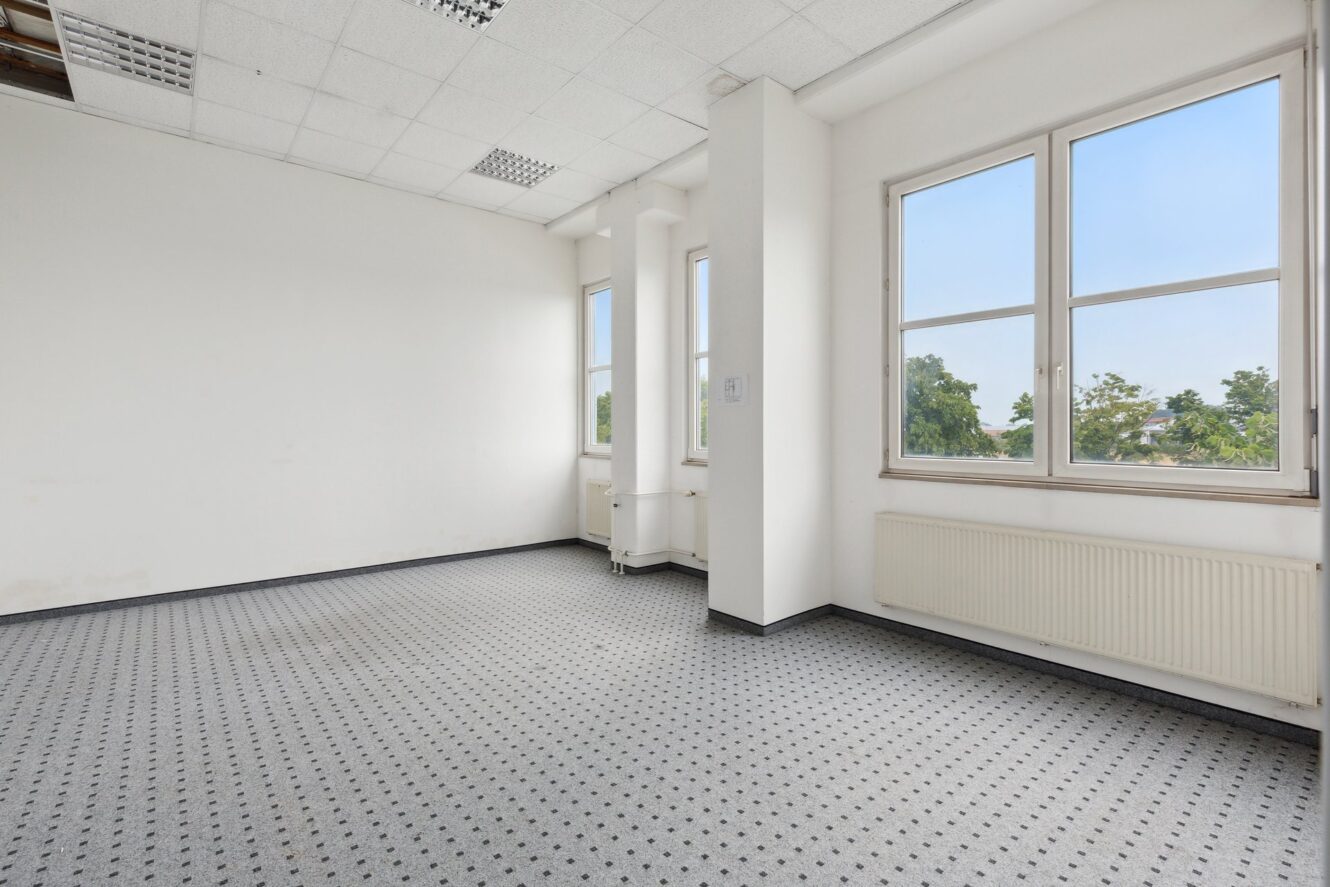 Leerer Büroraum mit weißen Wänden, gemustertem grauen Teppich, großen Fenstern, die viel Tageslicht hereinlassen, und einem Heizkörper unter den Fenstern.