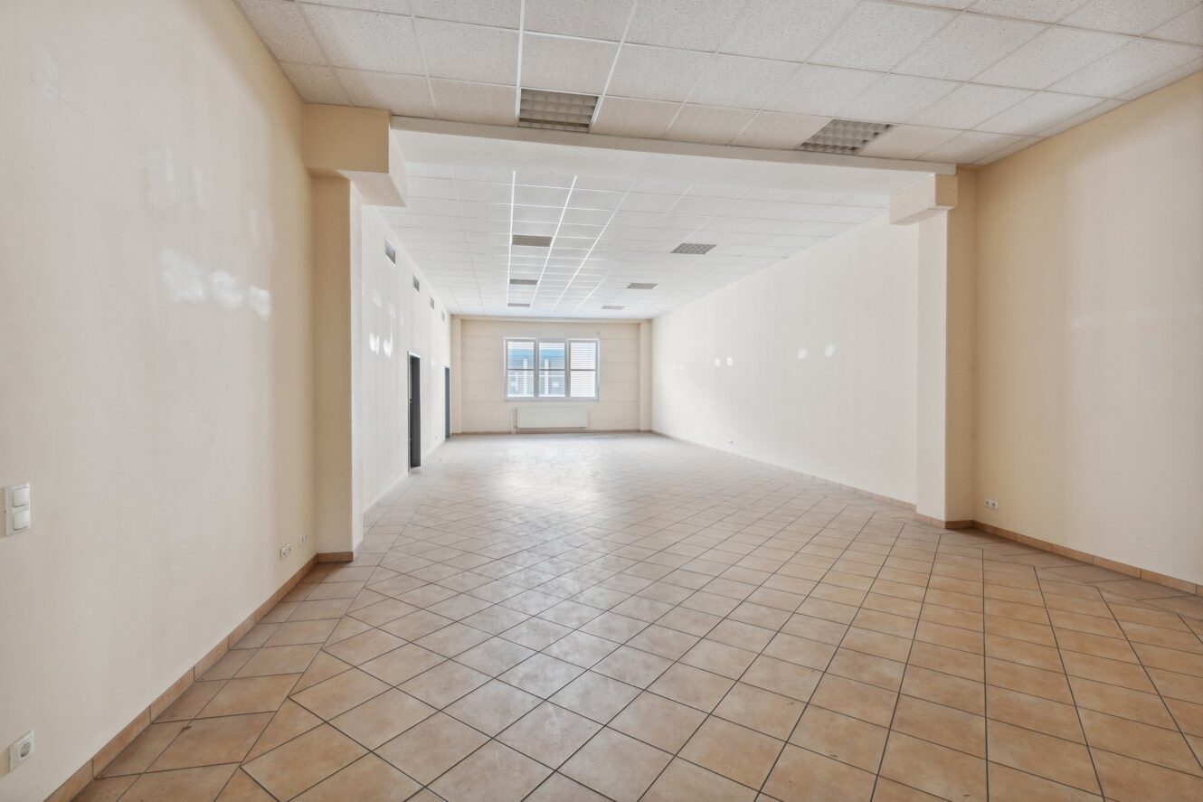 Leeres, unmöbliertes Zimmer mit gefliestem Boden, beigen Wänden, abgehängter Decke mit Leuchtstoffröhren und einem großen Fenster an der Stirnseite, durch das Tageslicht einfällt.