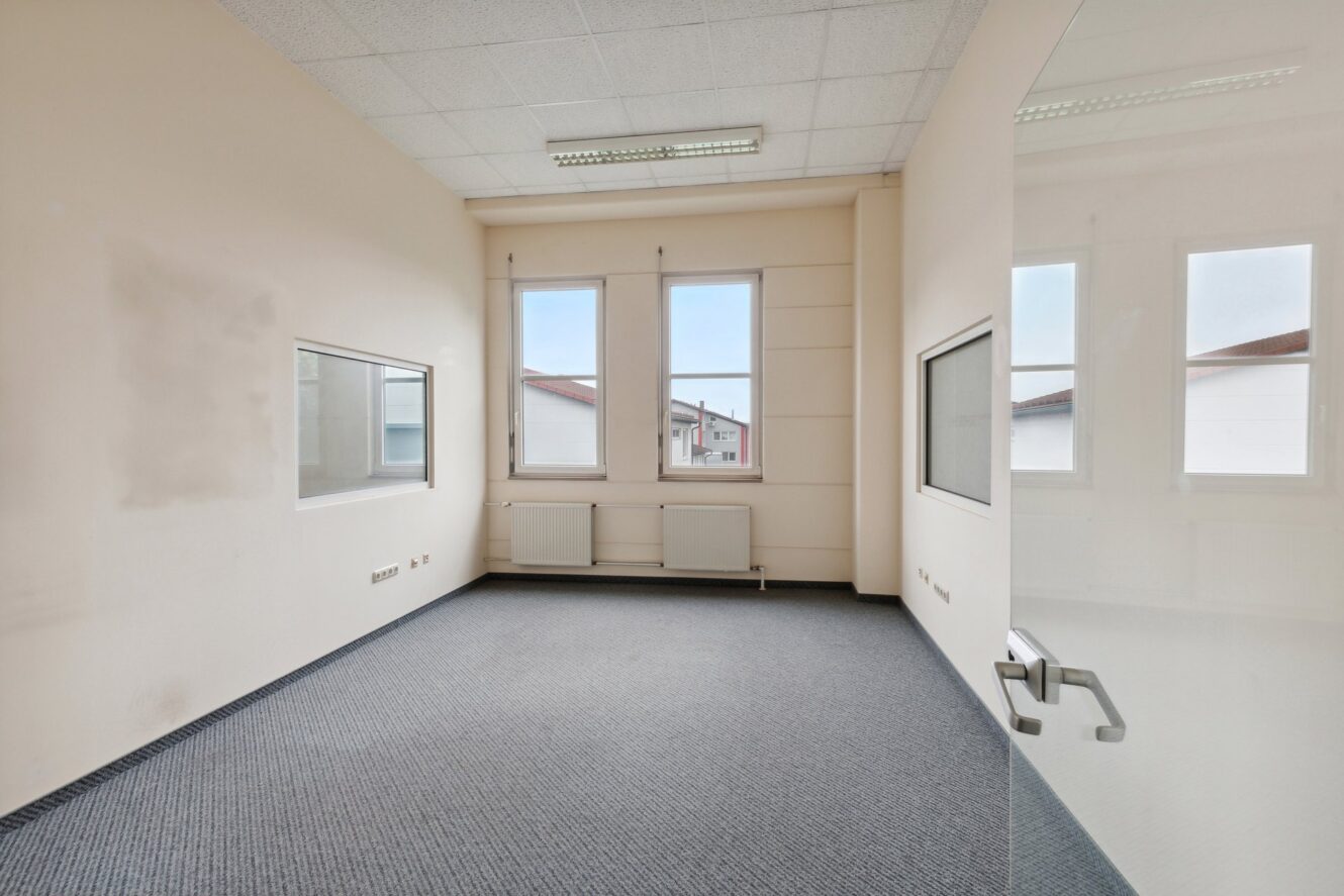 Leerer Büroraum mit weißen Wänden, zwei Fenstern, Wandheizkörpern, Teppichboden, fluoreszierenden Deckenleuchten und Glastür.