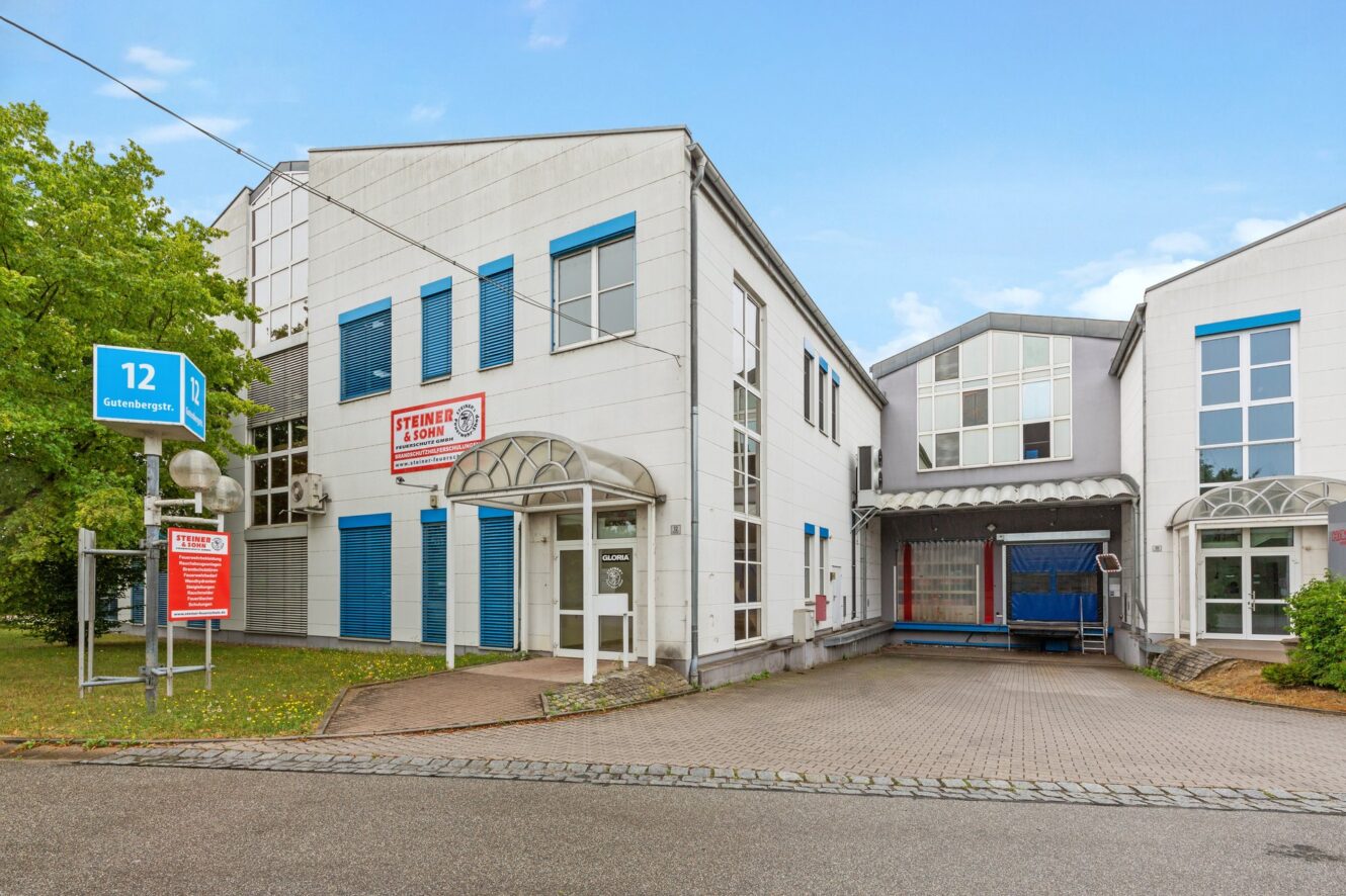 Zwei miteinander verbundene weiße Geschäftshäuser mit blauen Fensterläden; auf einem Schild steht Steiner Schule. Ein blaues Straßenschild trägt die Nummer 12. Bäume und eine gepflasterte Einfahrt sind zu sehen.