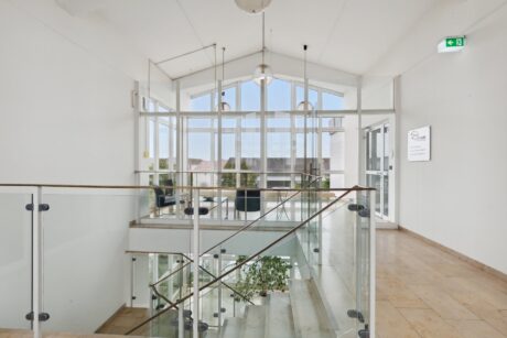 Moderne Innentreppe mit Glasgeländer, beigem Fliesenboden, großen Fenstern und Sitzecke; natürliches Licht erfüllt den Raum.