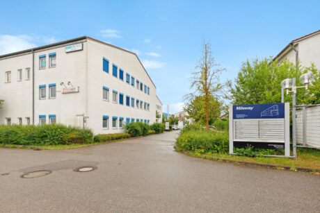 Ein weißes Bürogebäude mit blauen Fensterrahmen steht neben einer Einfahrt, mit einem großen Telefonbuchschild im Vordergrund und Grünflächen um das Grundstück herum.