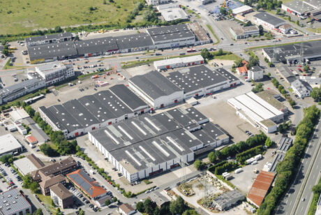 Luftaufnahme eines Industriekomplexes mit mehreren großen Lagerhallen und Gebäuden, umgeben von Straßen, Parkplätzen und einigen Grünflächen.