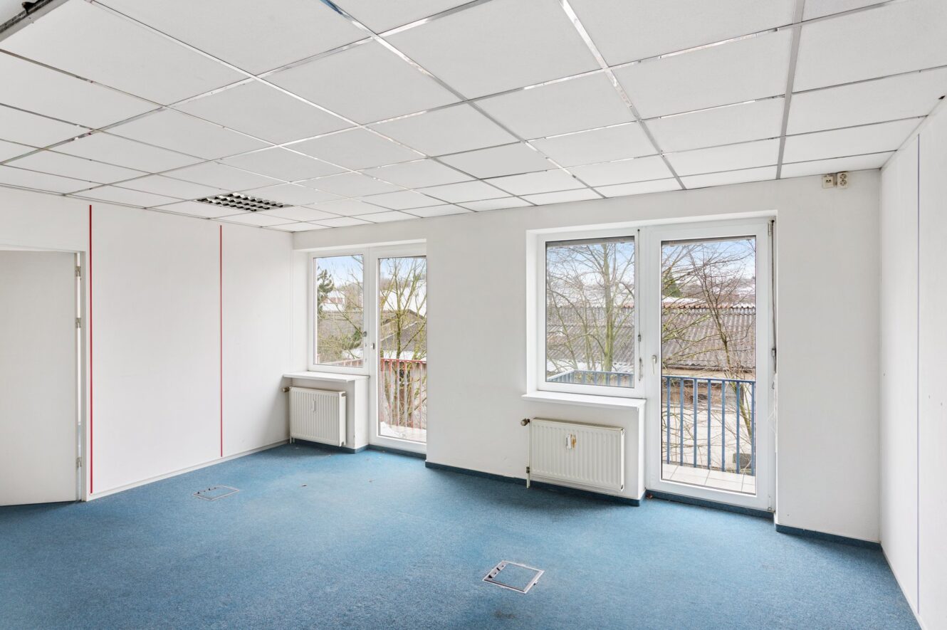 Ein leerer Büroraum mit blauem Teppich, weißen Wänden, einer abgehängten Decke mit Leuchtstoffröhren, zwei großen Fenstern und einer Tür auf der linken Seite.
