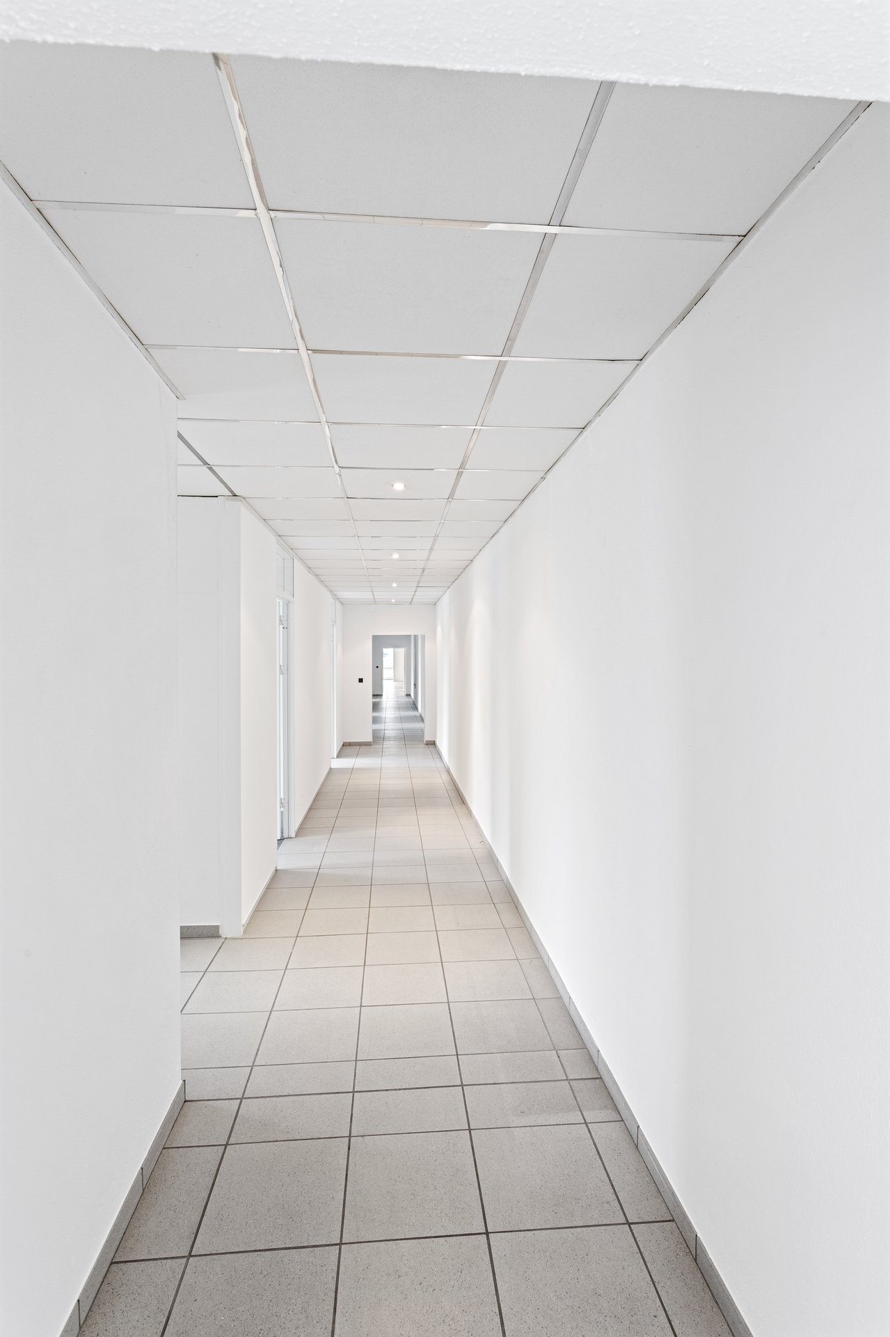 Ein langer, leerer Flur mit weißen Wänden, gefliestem Boden und einer abgehängten Decke mit Leuchtstoffröhren.