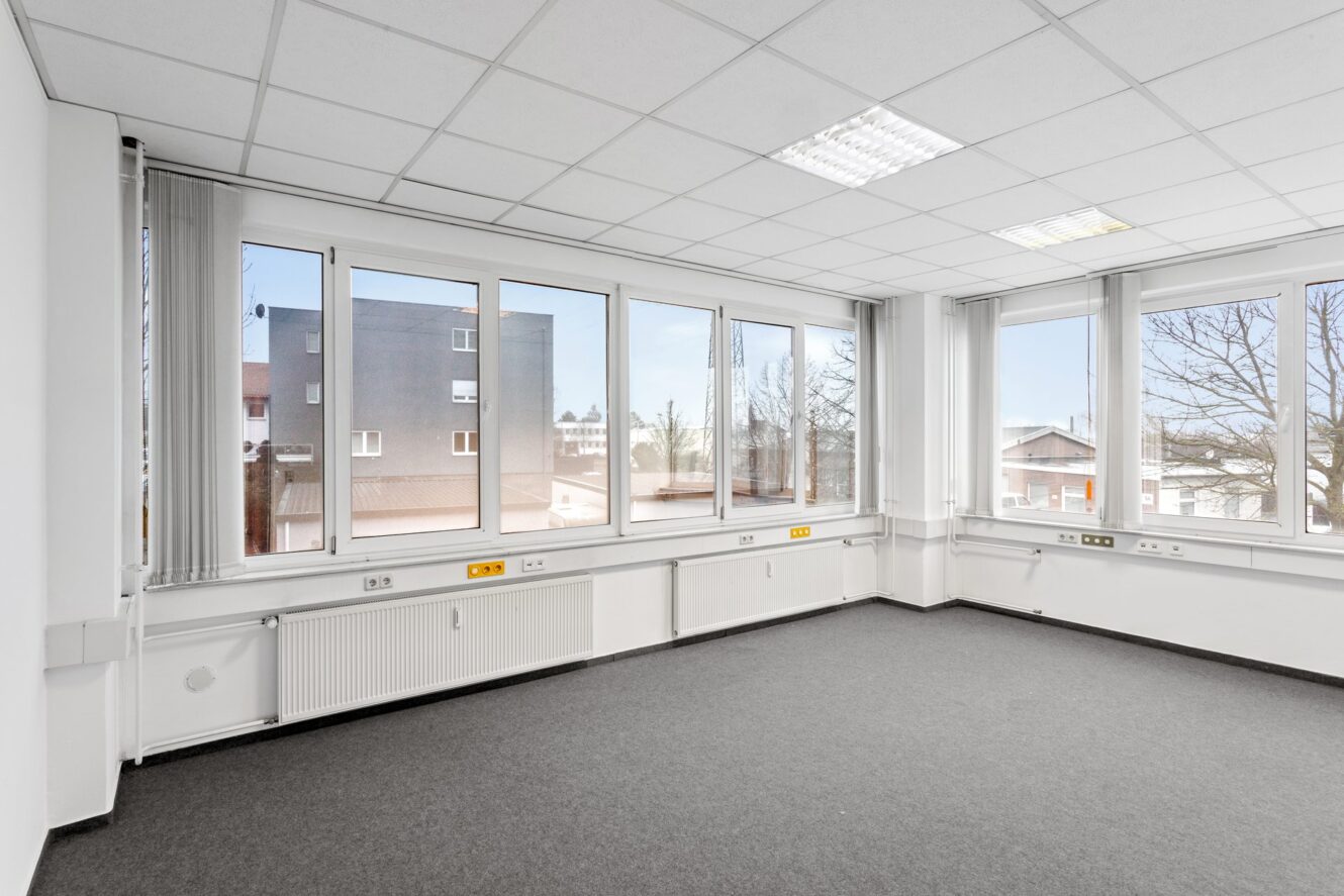 Leerer Büroraum mit großen Fenstern, weißen Wänden, grauem Teppich, Heizkörpern und Deckenleuchten; Blick auf Gebäude und Bäume draußen.