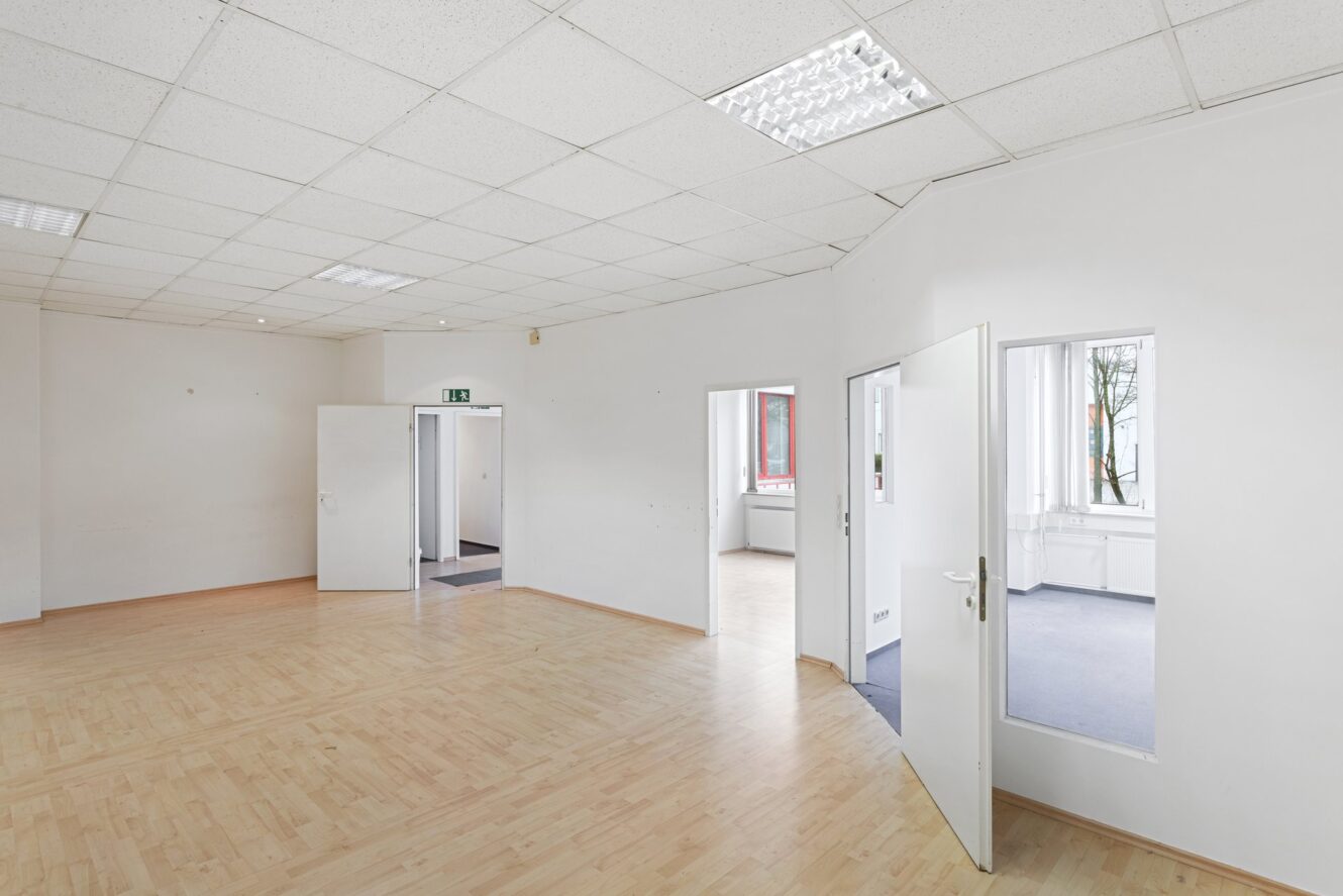 Leere Büroräume mit hellem Holzfußboden, weißen Wänden, offenen Türen, die zu anderen Räumen führen, und an der Decke angebrachten Leuchtstoffröhren.