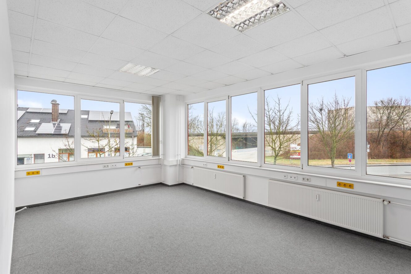 Leerer Büroraum mit weißen Wänden, grauem Teppich, großen Fenstern auf zwei Seiten und Deckenleuchten, die den Blick auf Gebäude und Bäume draußen freigeben.