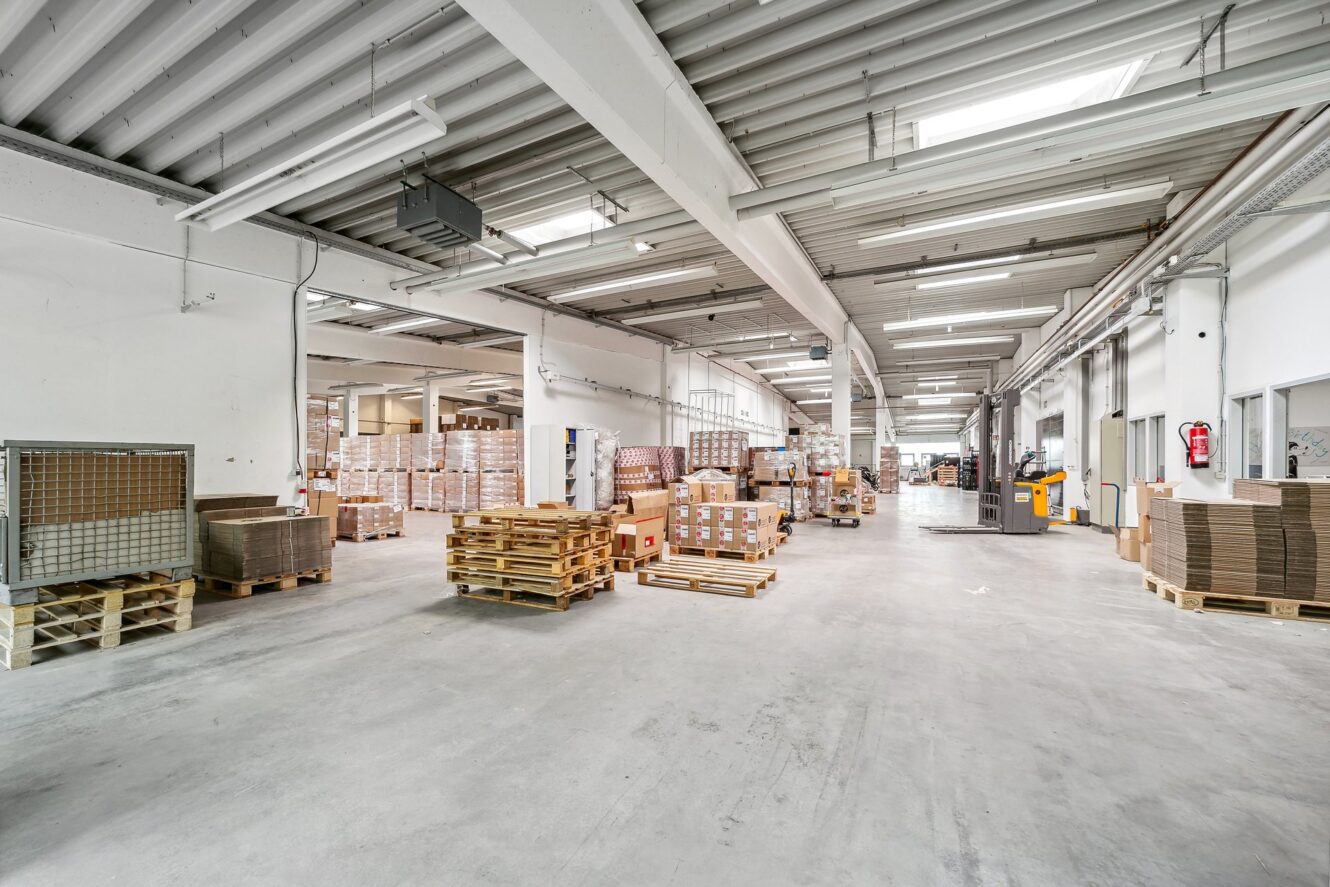 Geräumige Lagerhalle mit gestapelten Paletten, Kartons und Industriebeleuchtung, mit sauberem Betonboden und hoher Decke.