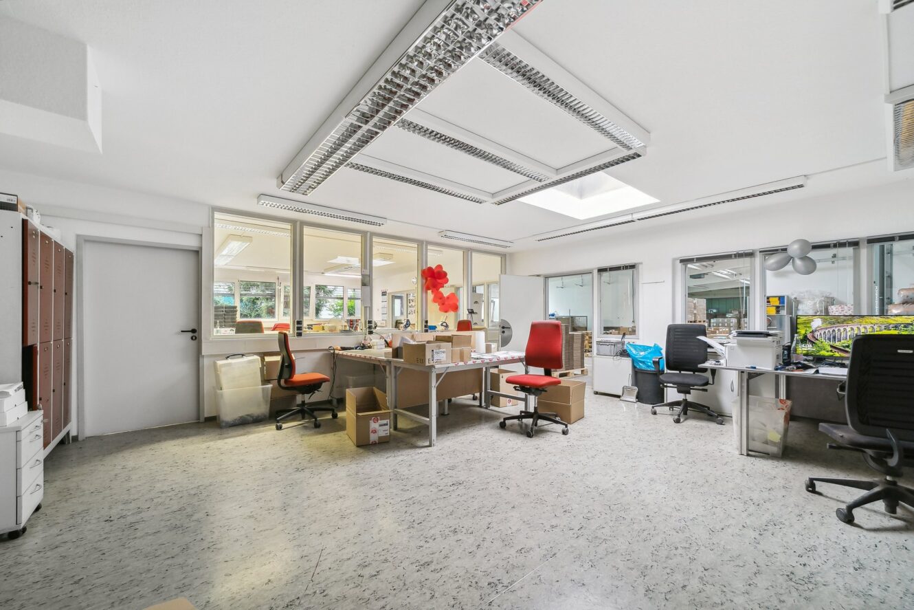 Heller Büroraum mit mehreren Schreibtischen, Stühlen, Computern und verstreuten Kisten; große Fenster und Leuchtstoffröhren erhellen den Raum.