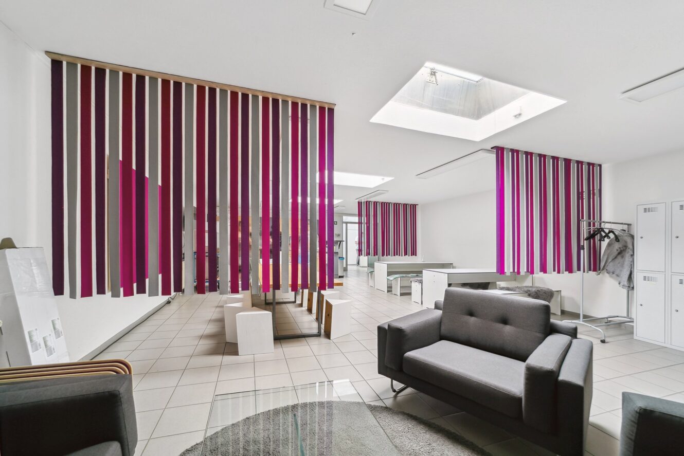 Moderner Büroraum mit gefliesten Böden, grauen Sitzmöbeln, Schließfächern und vertikal gestreiften Trennwänden in Lila-, Rosa- und Rottönen, mit natürlichem Licht durch Oberlichter.