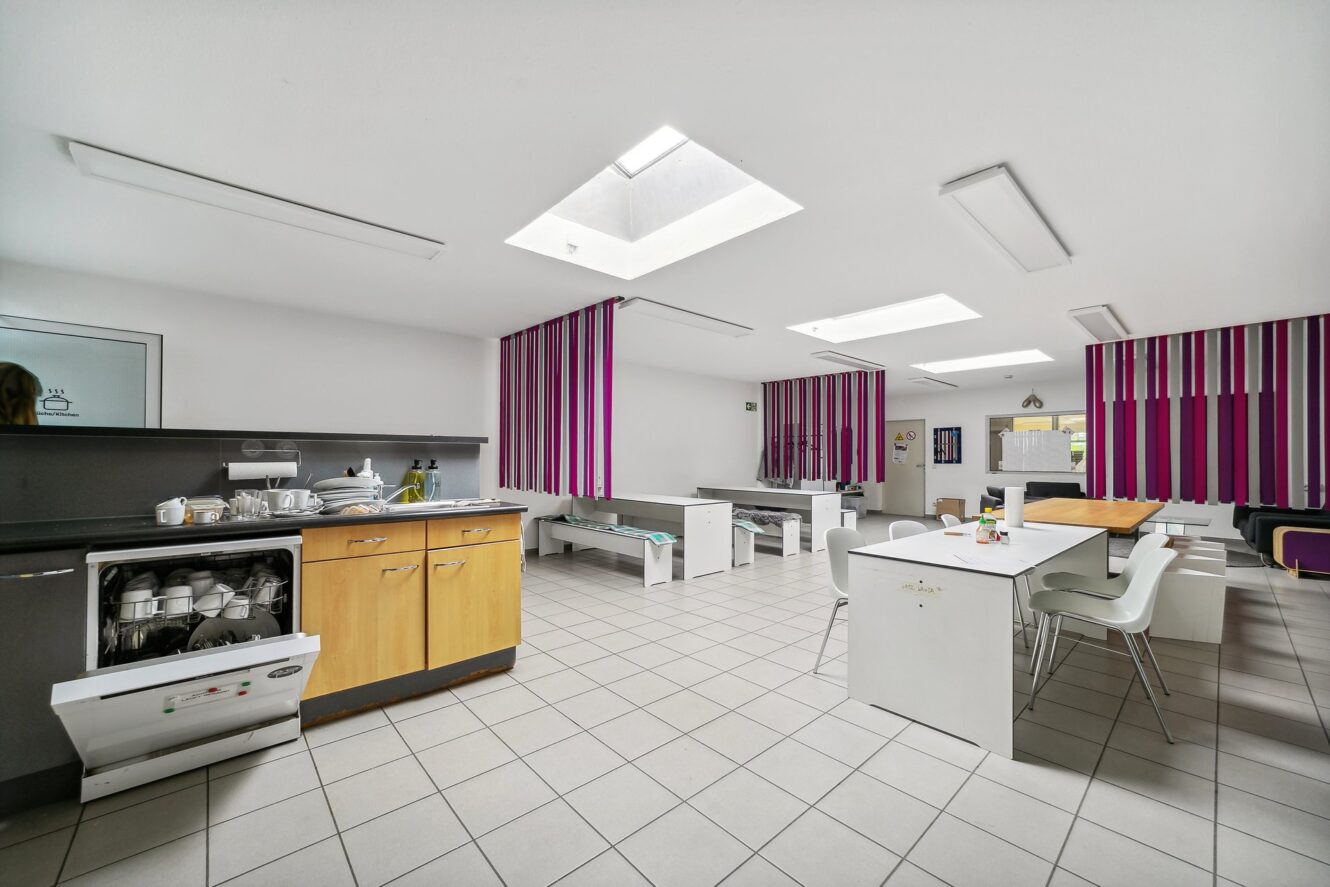 Moderne Gemeinschaftsküche mit offenem Geschirrspüler, Esstisch aus Holz, weißen Stühlen, gefliestem Boden und lila gestreiften Wandpaneelen unter Oberlichtern.