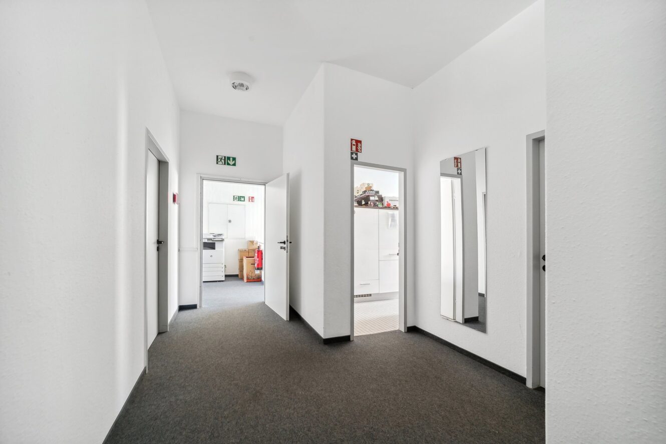 Ein sauberer, weißer Flur mit grauem Teppich, vier offenen Türen, zwei Wandspiegeln und einem Büro mit sichtbaren Möbeln am Ende.