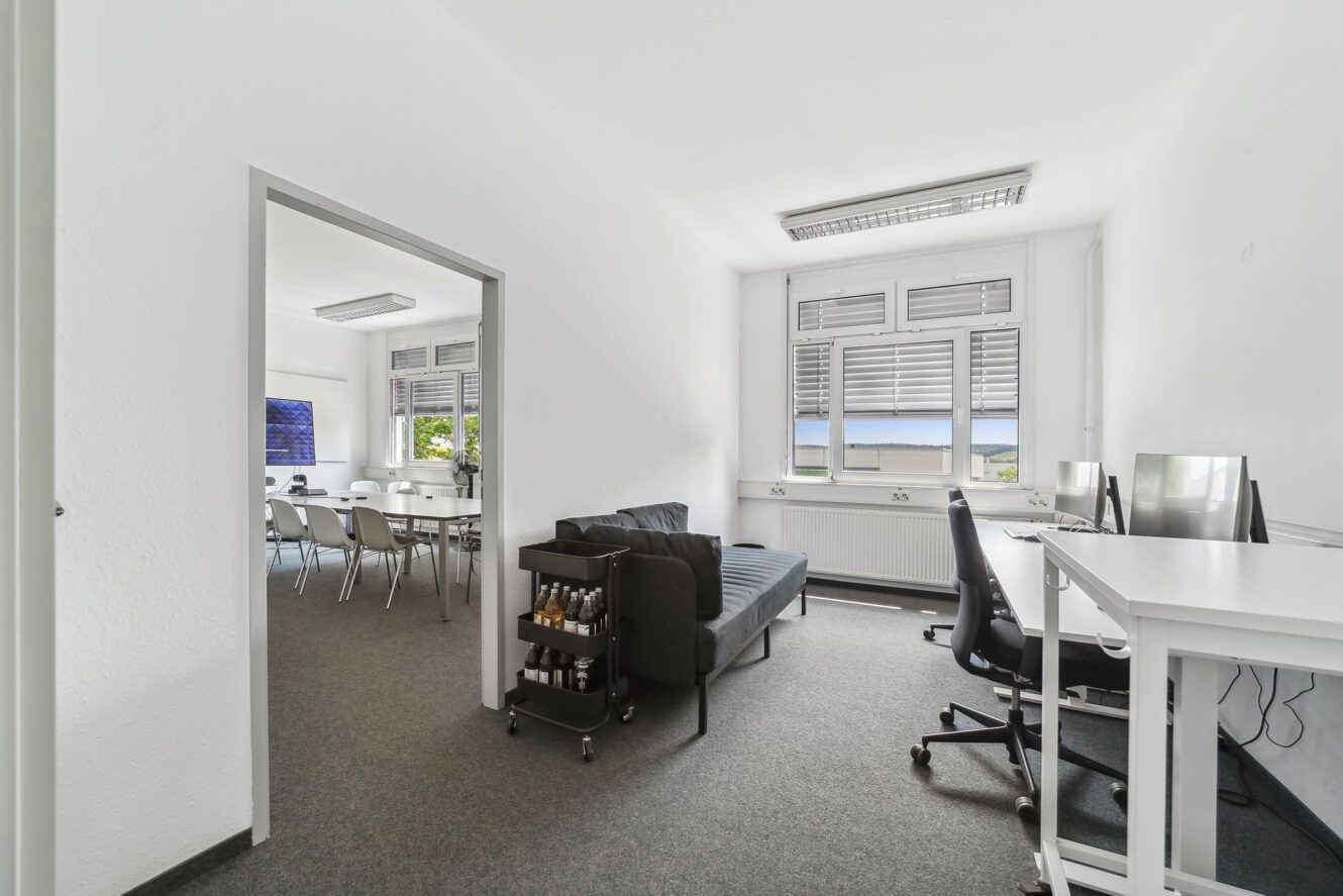 Moderner Büroraum mit weißen Wänden, zwei Schreibtischen, Bürostühlen, einem schwarzen Sofa, einem Getränkewagen und einem angrenzenden Konferenzraum mit einem Besprechungstisch und Stühlen.