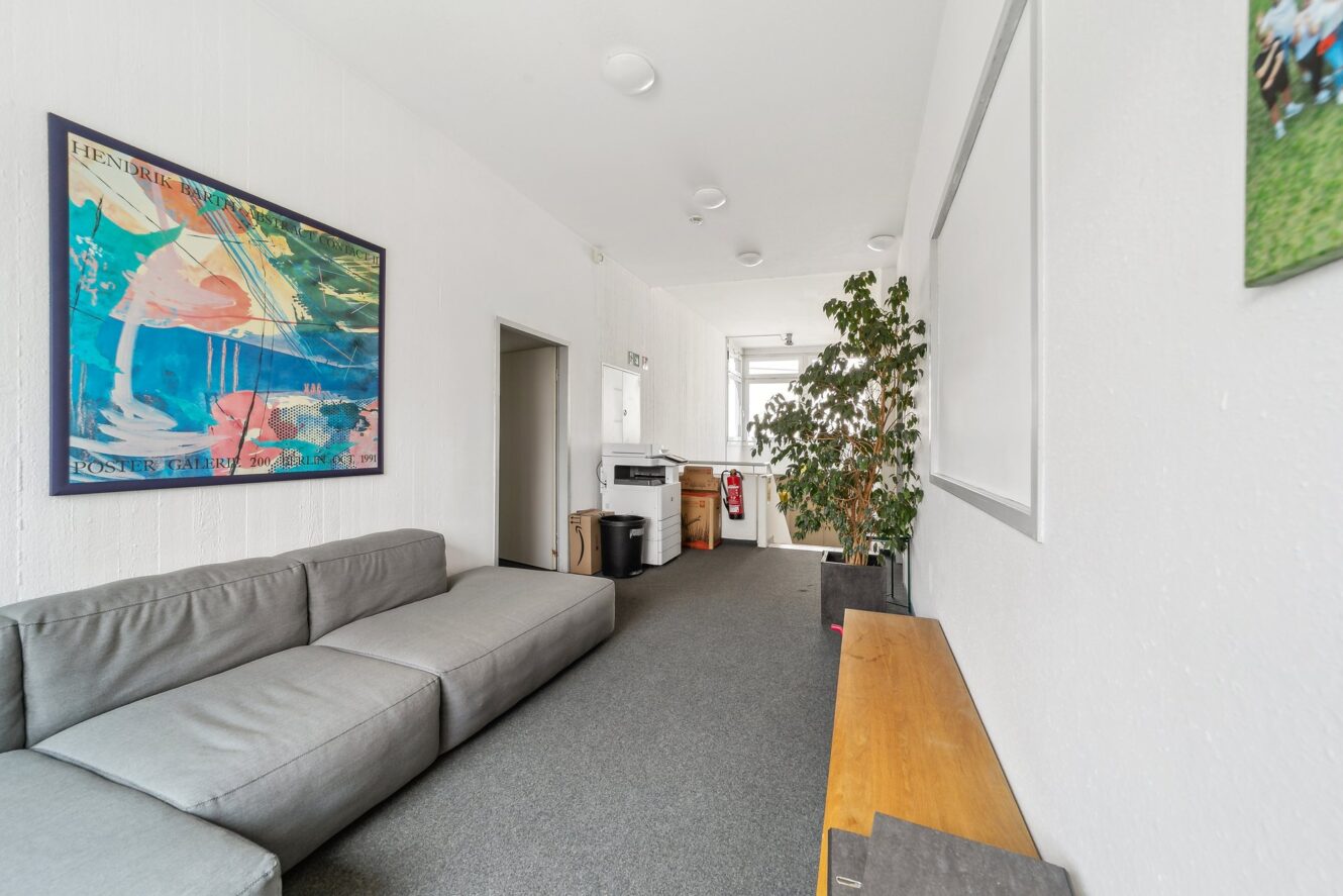 Ein moderner Lounge-Bereich mit einem grauen Sektionssofa, Wandkunst, einer Holzbank, einer Topfpflanze und einer Küchenzeile im Hintergrund.