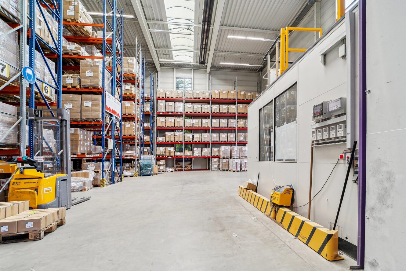 Ein großes Lager mit hohen Regalen voller Kisten, einem gelben Hubwagen und einem Kontrollraum mit Schalttafeln auf der rechten Seite.